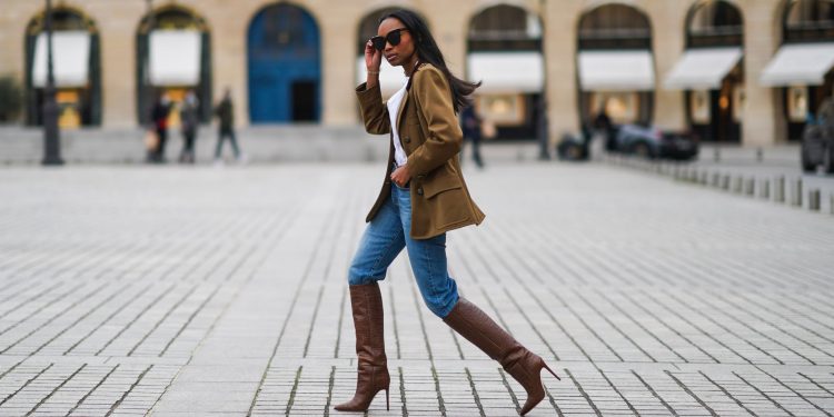 PARIS, FRANCE - JANUARY 26: Emilie Joseph wears sunglasses, a khaki military jacket from Saint-Laurent YSL, a white shirt / blouse from Zara with ruffled collar, blue denim jeans pants from Levi's, brown high heeled knee-high pointy leather crocodile pattern boots from Paris Texas, on January 26, 2021 in Paris, France. (Photo by Edward Berthelot/Getty Images)